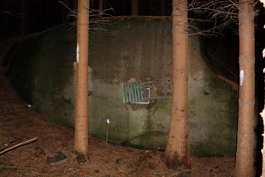 Židlička a kus polystyrenu, přivázaná uprostřed lesa řetězem ke skále.